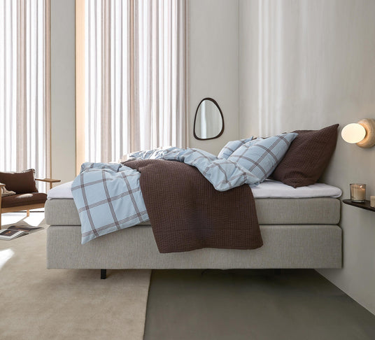 Lyst soveværelse med beige polstret seng, blåternet sengetøj med brune puder og plaid, væglampe, spejl og tændt duftlys på lille hylde, blødt dagslys gennem lette gardiner.