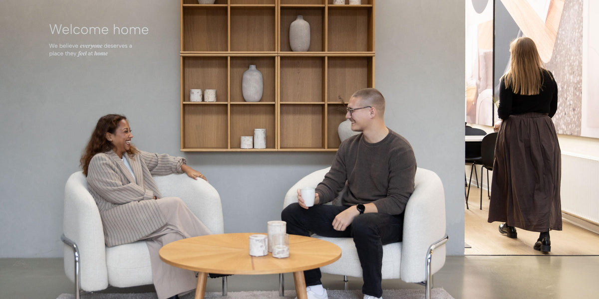 Velkommen hjem – vi mener, at alle fortjener et sted, hvor de føler sig hjemme; to personer sidder i hvide lænestole ved et rundt sofabord med lys og keramik foran en træreol i et lyst showroom.