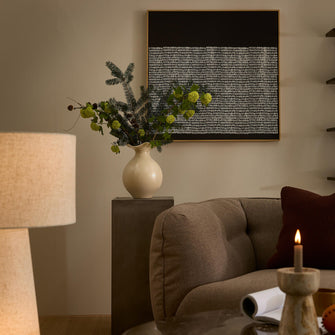 Hyggeligt hjørne i stuen med beige sofa, hvid keramikvase med grønne grene på sokkel, bordlampe og tændt lys, abstrakt sort‑hvidt vægbillede.