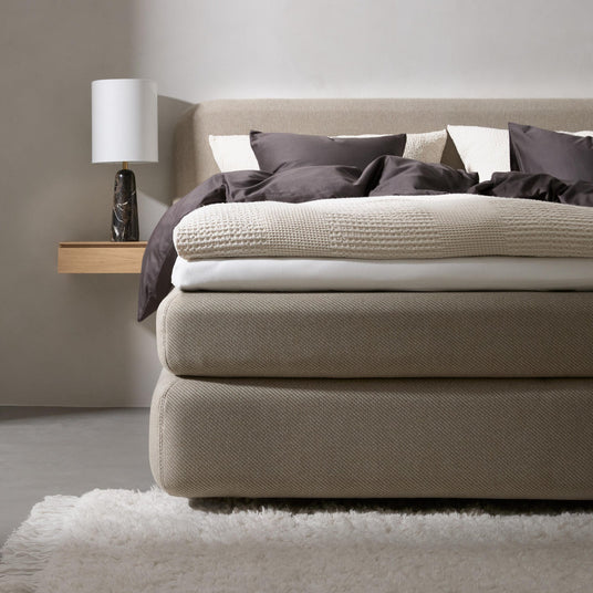 Beige polstret seng redt med mørkegrå og cremefarvede tekstiler, svævende træhylde med bordlampe og blød hvid tæppe i et minimalistisk soveværelse.