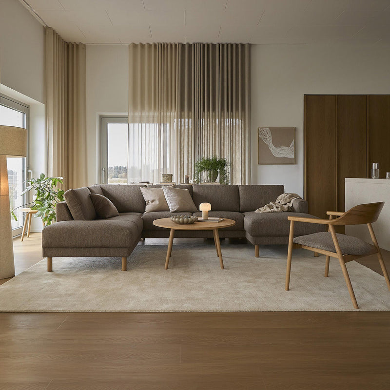 Lys og varm stue med beige gardiner, brun modulsofa, rundt sofabord i træ med tændt lys og stort lyst tæppe.