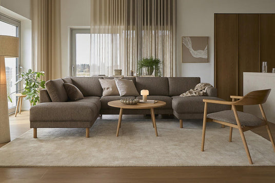 Lys og varm stue med beige gardiner, brun modulsofa, rundt sofabord i træ med tændt lys og stort lyst tæppe.