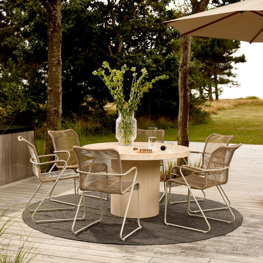 Rundt lyst træbord på terrasse med fire flettede udestole, stor glasvase med grønne stilke, glas og lille skakspil på bordet, gråt rundt tæppe og parasol i dagslys.