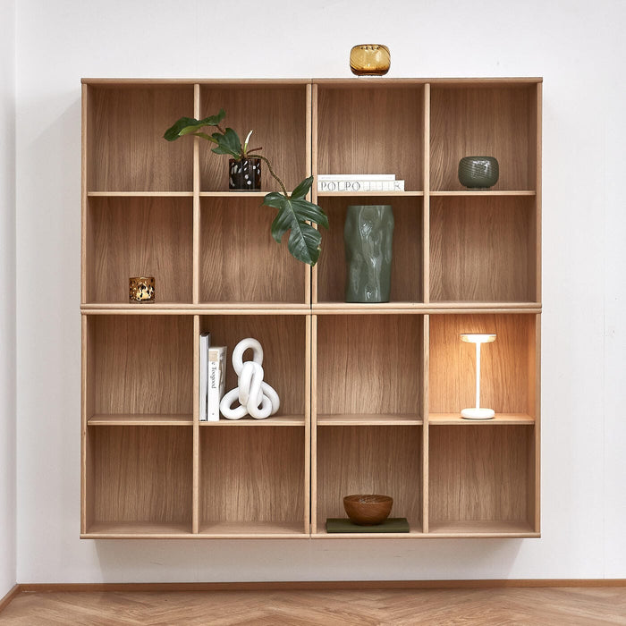 Vægmonteret reol i træ med bøger, plante, grøn vase, små skåle og en hvid trådløs bordlampe; en bog med titlen POLPO ses.