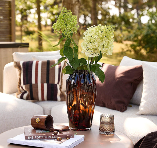 Ravfarvet glasvase med hortensia på et udendørs sofabord; ved siden af står et struktureret glas, en træ-terningebæger med terninger og bøger med titlen THE POTTED GARDEN; lyse puder i baggrunden.
