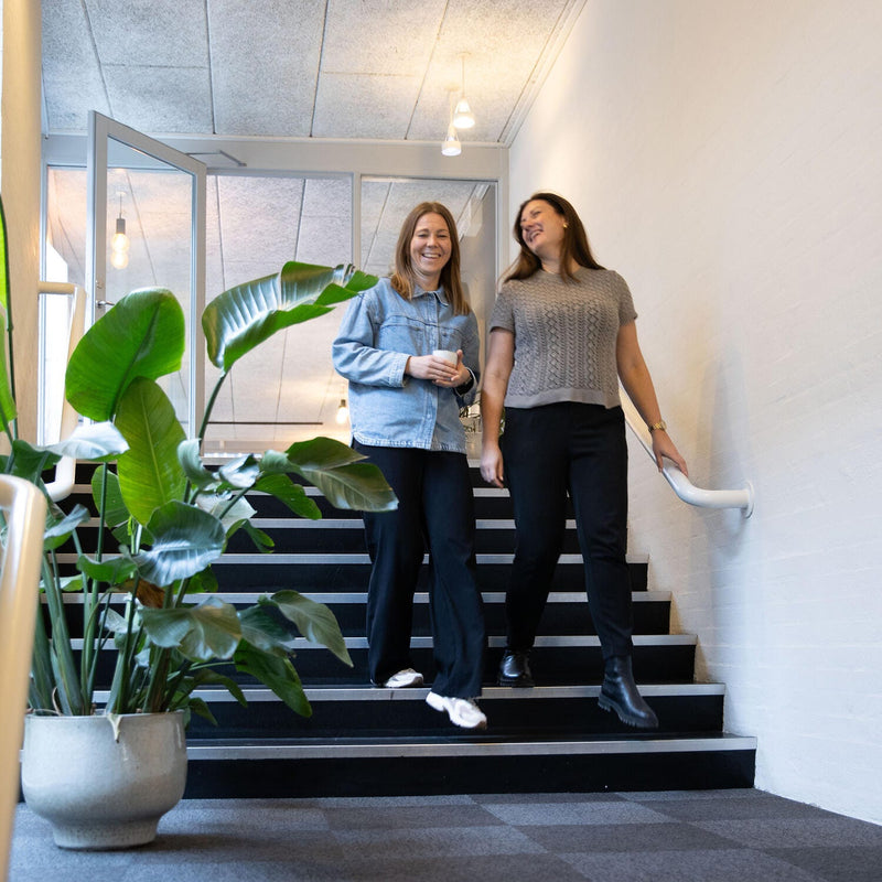 To personer går ned ad en indendørs trappe i en lys gang; den ene holder et krus og den anden i gelænderet; stor potteplante og loftslamper i baggrunden.