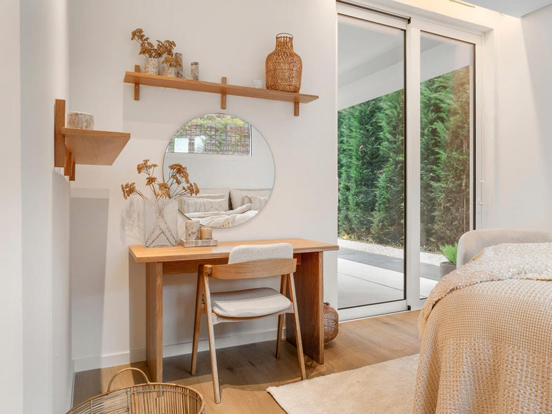 Lyst soveværelseshjørne i skandinavisk stil med træskrivebord, rundt spejl, beige tekstiler, kurv og tørrede planter ved store skydedøre.