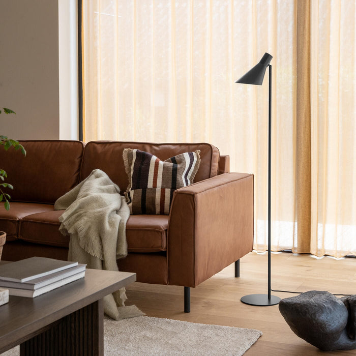 Brun lædersofa med stribet pude og lys plaid, sort gulvlampe og træbord i stue med beige gardiner i dagslys.