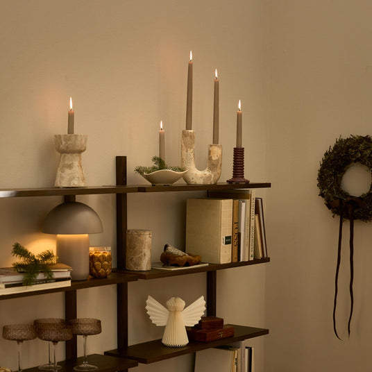 Mørk træreol med tændte lys i lysestager, en varmglødende bordlampe, bøger og glas; en grøn krans hænger på væggen.