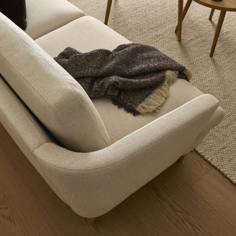 Lys beige sofa i stuen med brunmeleret plaid og lyst uldtæppe på trægulv.