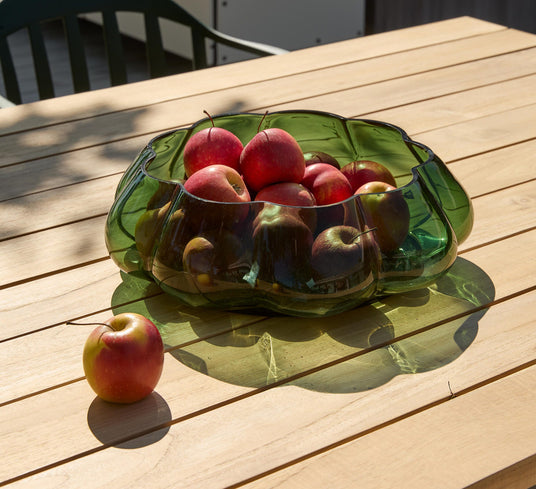 Grøn glasskål fyldt med røde æbler på et lyst havebord i solskin.