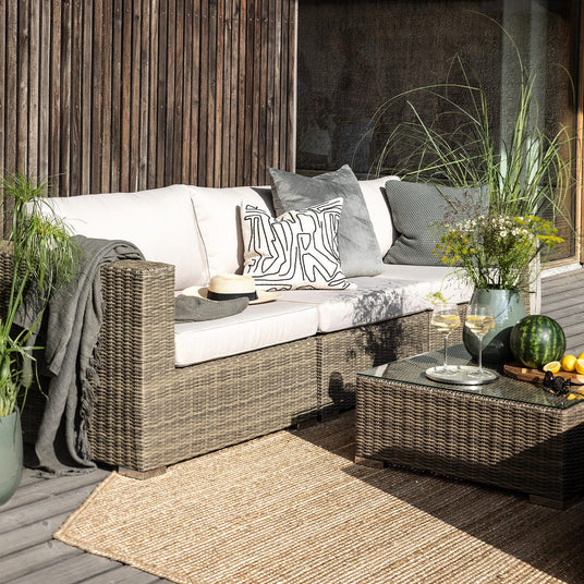 Terrasse med fletsofa, puder og plaid, lavt bord med to vinglas, vandmelon og citroner, omgivet af potteplanter i sollys.