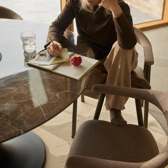 Person, der skriver i en notesbog ved et rundt marmorbord; et glas og et rødt æble ligger på bordet i blødt dagslys i hjemmemiljø.