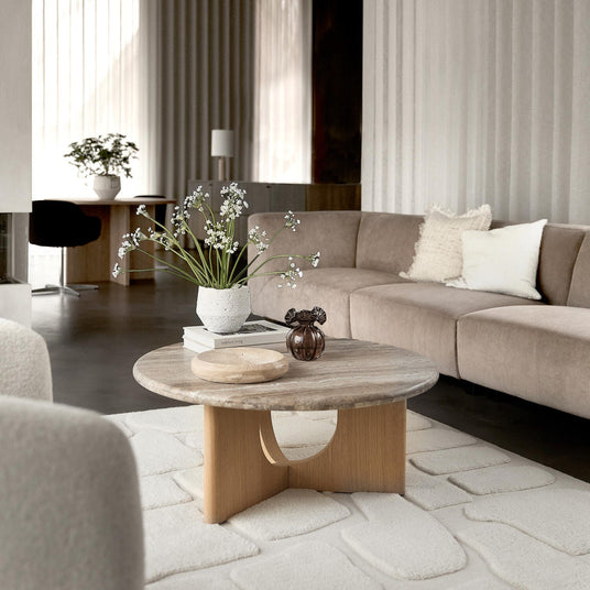 Lys skandinavisk stue med beige modulsofa, rundt sofabord i træ og sten med vase med hvide blomster, lille brun glasdekoration og skål på bøger ("lifestyle living"), lyse gardiner og tekstureret hvidt tæppe.