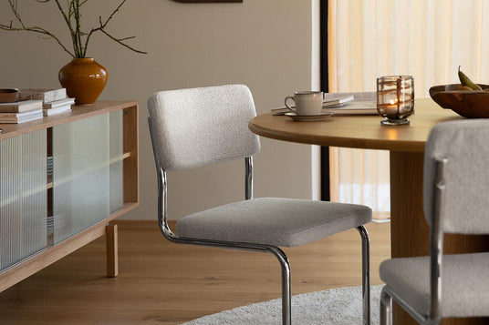 Lys grå polstret cantilever-stol ved rundt spisebord i træ i en skandinavisk spisestue med ribbet glasskænk, pendel, vase med grene og tændt fyrfadslys på et neutralt rundt tæppe.