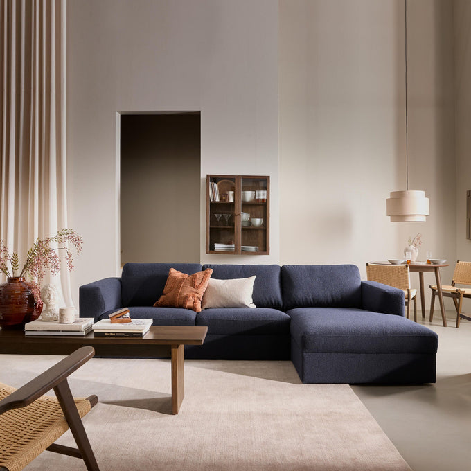 Beige stue i blødt dagslys med mørkeblå hjørnesofa, træsofabor, puder, tæppe, gardiner, vægskab og en lille spiseplads med fletstole og beige pendel.