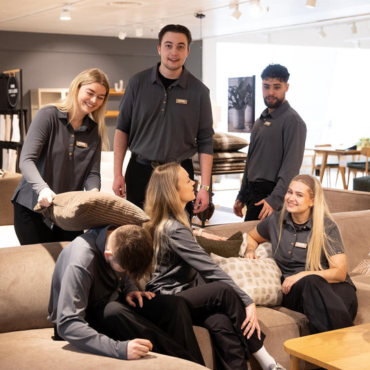 Gruppe af ILVA‑medarbejdere i en møbelbutik omkring en beige hjørnesofa, nogle leger med puder; pendellamper, træbord og stor hvid vase med tørrede planter i forgrunden.