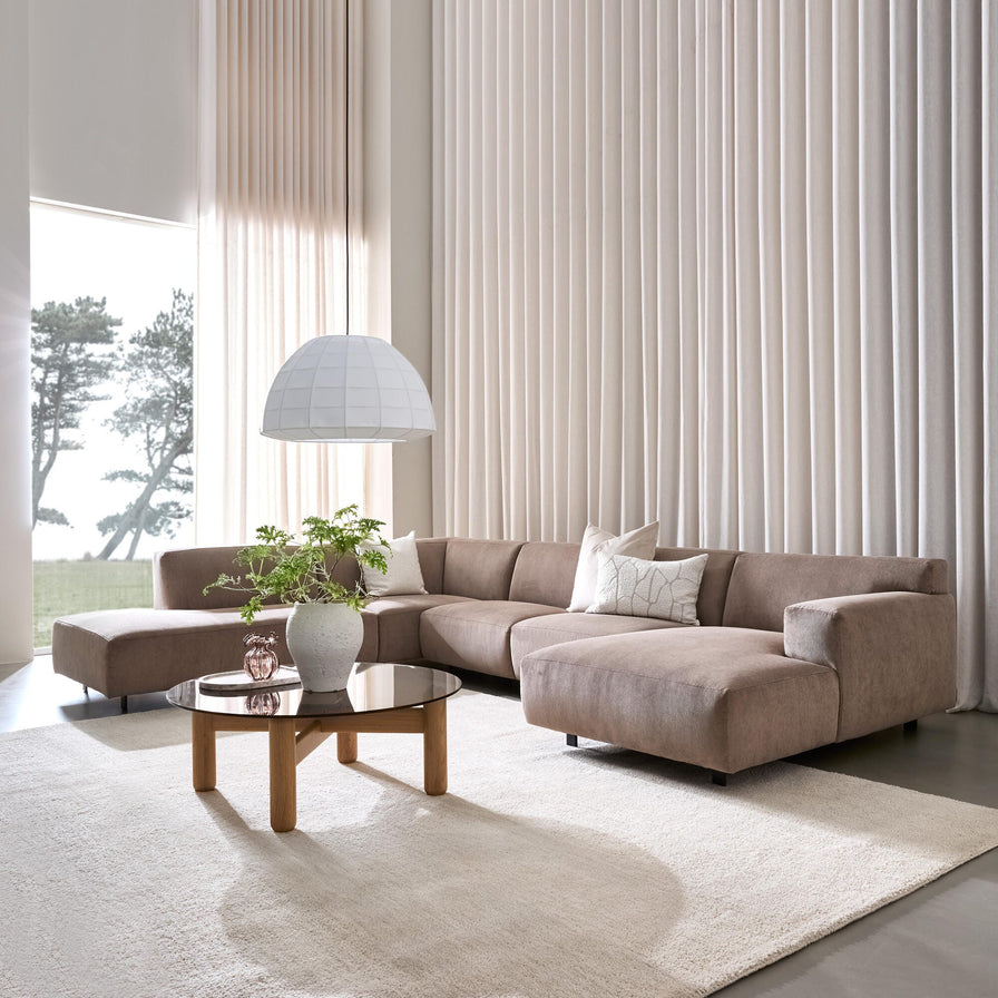 Lys stue med beige gardiner, brun modulsofa i hjørne, rundt sofabord i træ med glas og grøn plante, stor hvid pendellampe og blødt lyst tæppe i dagslys.
