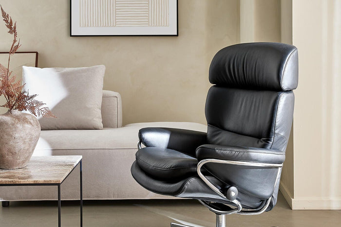 Minimalistisk stue med en sort læderlænestol på krom sokkel ved siden af en lys beige sofa, solstriber på gulvet, marmorbord med vase og tørrede grene samt indramet abstrakt linjekunst på væggen.