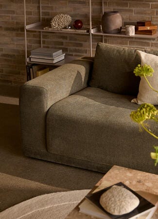 Stue med grå stofsofa, mønstrede puder og ravfarvet vase med grønne blomster på sofabord; murstensvæg og hylder i varmt dagslys.