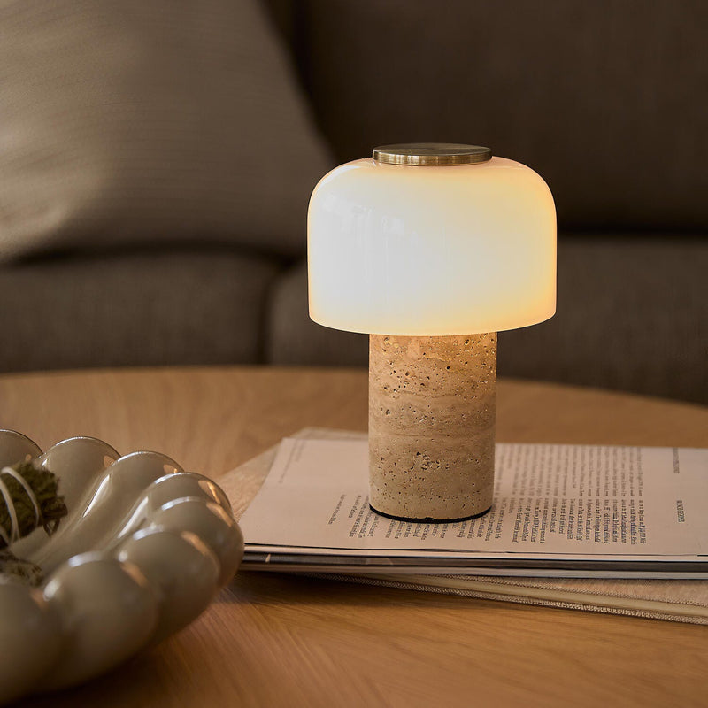 Lille bordlampe med travertinfod og opalglas, tændt på et sofabord med magasiner og skulpturelt fad i en hyggelig stue.