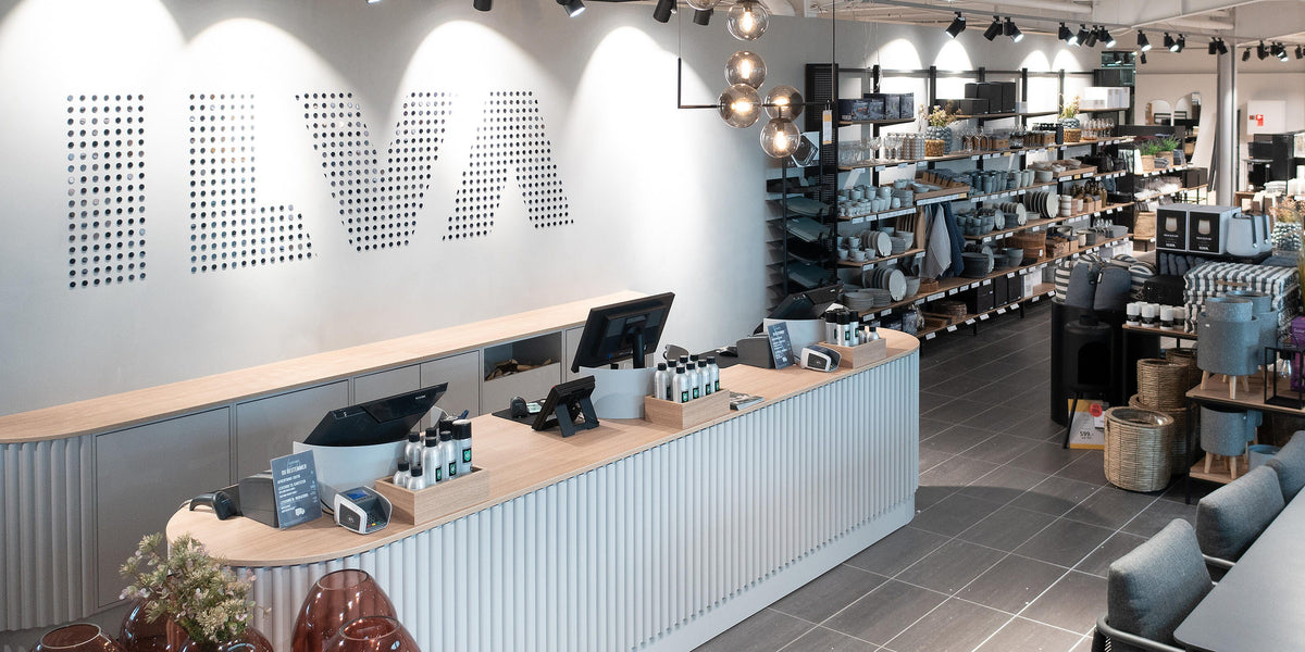 Moderne boligbutik med kasse, ILVA-logo på væggen og hylder med service, kurve og lamper; pris 99,- ses.