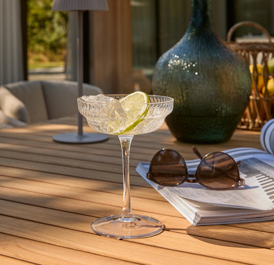 Cocktailglas med is og lime på et solbeskinnet træbord udendørs, med solbriller og magasin ved siden af.