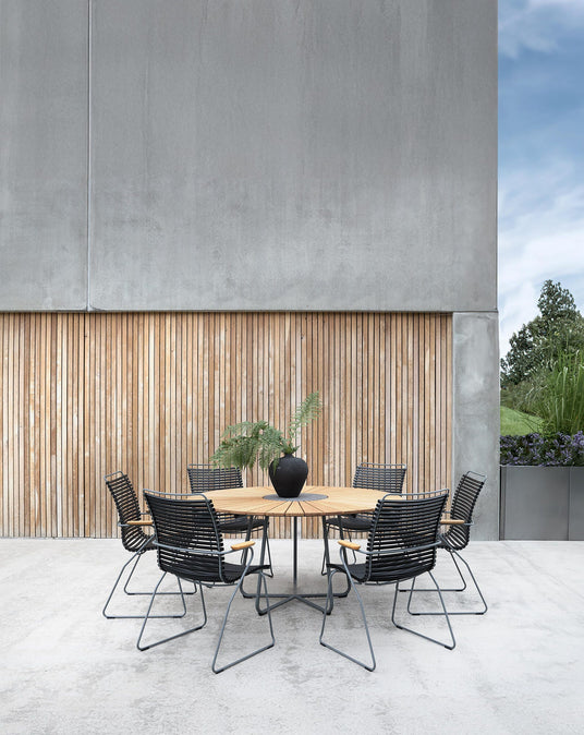 Rundt udebord i træ med seks sorte stole på en betonterrasse, sort vase med grønne grene som centerpiece foran en væg med lodrette trælister.