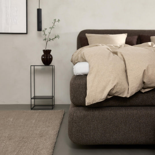 Minimalistisk soveværelse med brun polstret seng, beige linned sengetøj, sort metal-natbord med vase og gren, hængende pendellampe og neutral løber.
