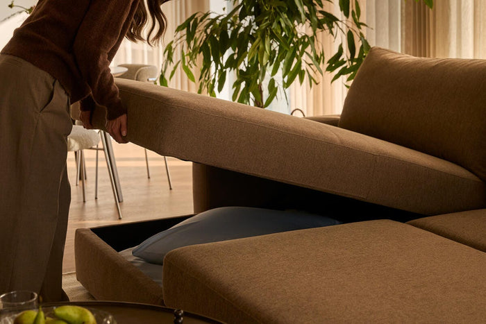 Brun modulsofa med opbevaring i sædet åbnes af en person; sofabord med pærer og spil i en lys stue med grønne planter.