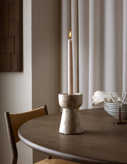 Tændt slankt stearinlys i stenlysestage på rundt spisebord, med stakke af skåle, serviet og cocktailglas ved siden af og neutrale gardiner i baggrunden.