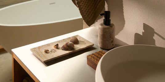 Nærbillede af et minimalistisk badeværelse med rund håndvask, stensæbedispenser og bakke med strandskaller og smykker; fritstående badekar i baggrunden.