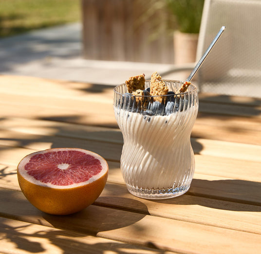 Yoghurt i riflet glas med blåbær og granola og en ske på et solbelyst træbord, ved siden af en halveret grapefrugt.