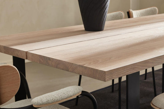 Nærbillede af et spisebord i lys eg med sorte ben, beige polstrede stole og en mørk vase.
