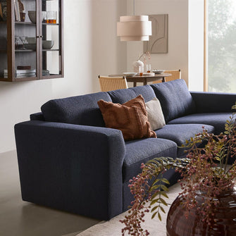 Mørkeblå hjørnesofa med rust- og beige pyntepuder i en lys stue; vase med grene i forgrunden, pendel og lille spiseplads i baggrunden.