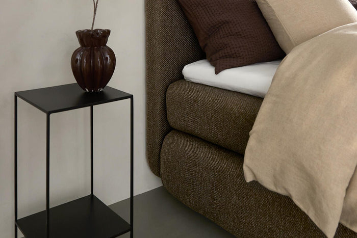 Nærbillede af soveværelse med brun polstret seng, beige og brune puder, sort metal-sidebord med brun vase og gren samt en hængende sort pendellampe.