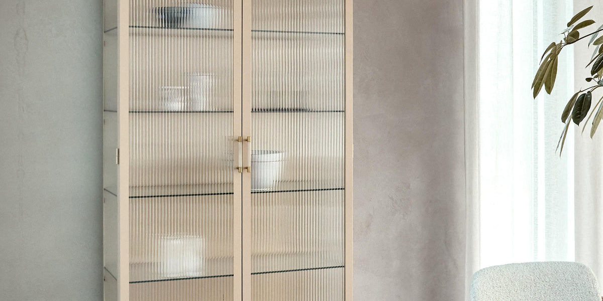 Beige vitrineskab med rillet glas og slanke ben i et lyst spisemiljø; rund træbordplade og let gardin i dagslys.