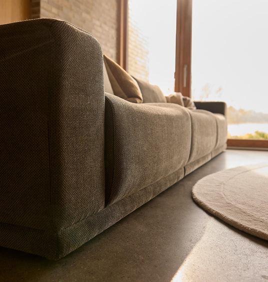 Nærbillede af beige stofsofa med puder i en solrig stue ved store vinduer, med lys rund måtte på betongulv.