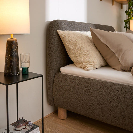 Nærbillede af en opredt seng med beige puder, strikket plaid og smalt natbord med mørk marmorlamp, glas og bøger i et varmt og hyggeligt soveværelse.