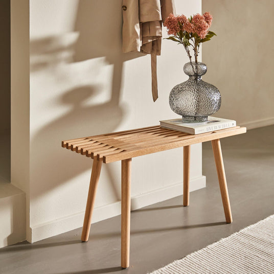Lys træbænk mod beige væg med struktureret glasvase med lyserøde blomster på en stak bøger; blødt dagslys og en hængt frakke kaster skygger.