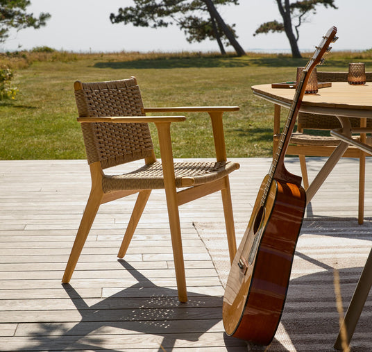 Træ- og fletstol på solrig terrasse ved rundt bord; en akustisk guitar læner sig mod bordet med udsigt til græs og træer.
