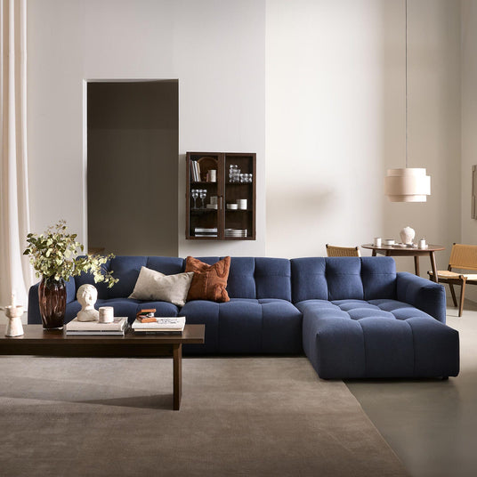 Stilrent stue-miljø med blå modulsofa, mørkt træbord med vase og bøger, beige gardiner og en pendel i en lys, neutral palet.