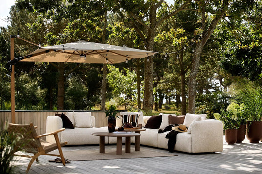 Udendørs lounge på træterrasse med lys modulsofa, runde sofaborde, parasol med lyskæde, puder og plaid, omgivet af træer og krukkeplanter i dagslys.