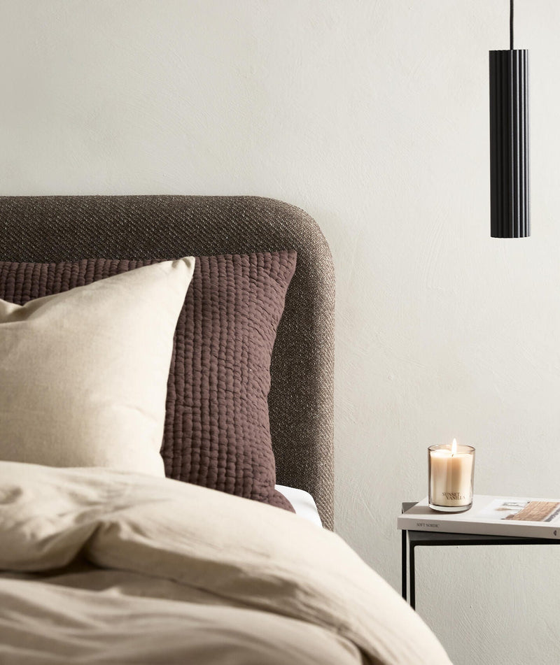 Neutralt soveværelse med beige sengetøj, brun quiltet pude, sort pendellampe og sidebord med tændt vaniljelys og bogen New Nordic.