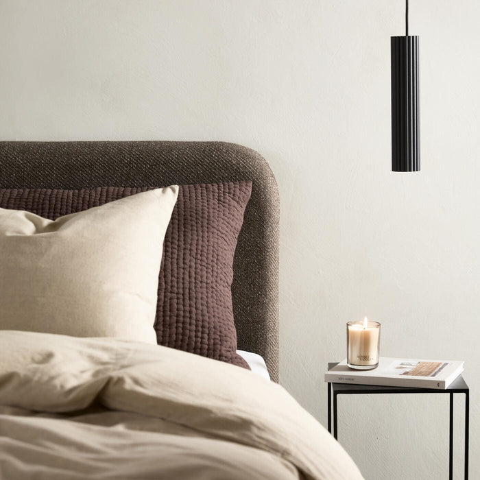 Neutralt soveværelse med beige sengetøj, brun quiltet pude, sort pendellampe og sidebord med tændt vaniljelys og bogen New Nordic.