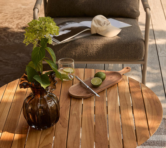 Rundt teakbord på terrasse med brun glasvase med hortensia, glas med lime, træbræt med halveret lime og en pen; i baggrunden en loungestol med åbent magasin og kasket.