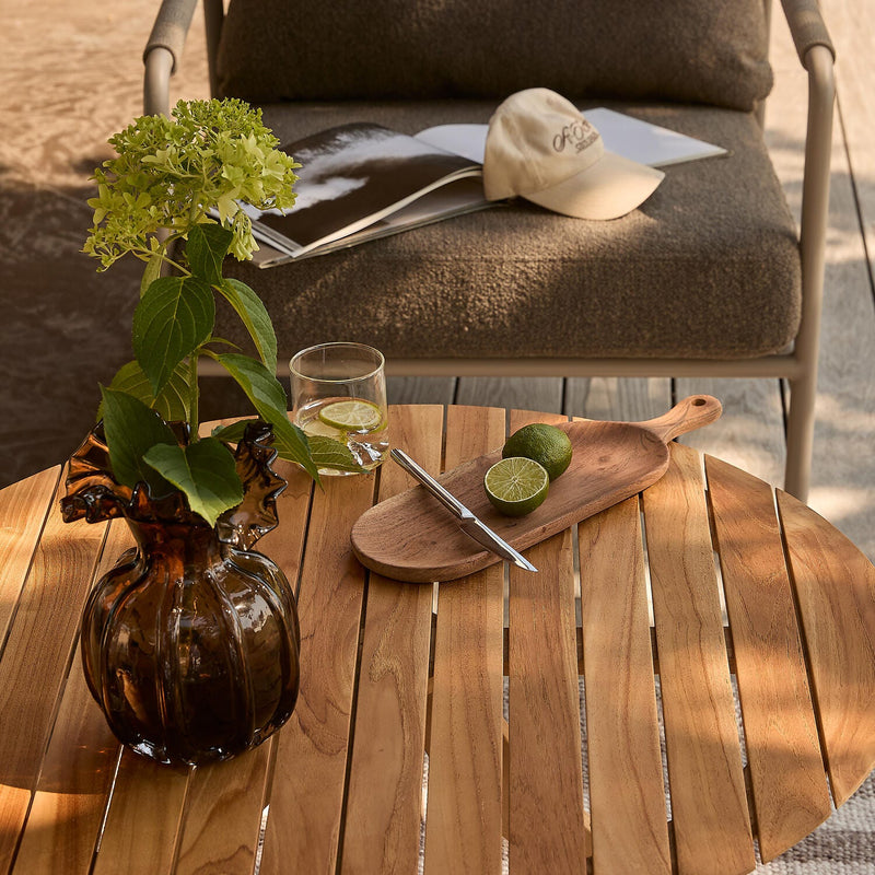 Rundt teakbord på terrasse med brun glasvase med hortensia, glas med lime, træbræt med halveret lime og en pen; i baggrunden en loungestol med åbent magasin og kasket.