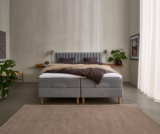 Minimalistisk soveværelse med grå polstret dobbeltseng, neutralt sengetøj i brune og beige nuancer, planter, væglamper, indramet kunst og lyse gardiner.