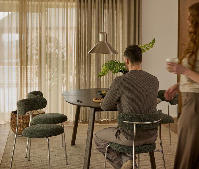 Spiseplads med rundt mørkt bord, grønpolstrede stole, pendellampe og grøn plante; en mand sidder, og en kvinde går forbi med et glas foran lyse gardiner.