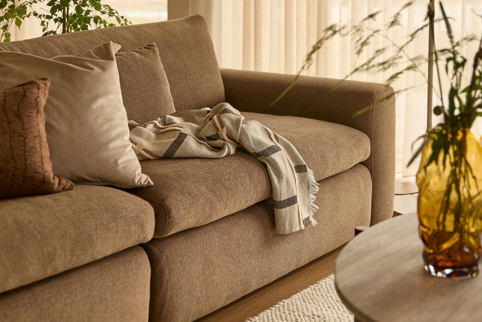 Beige-brun sofa med puder og en stribet plaid, foran en rund træbordplade med en ravfarvet vase; lette gardiner og grøn plante i blødt dagslys.